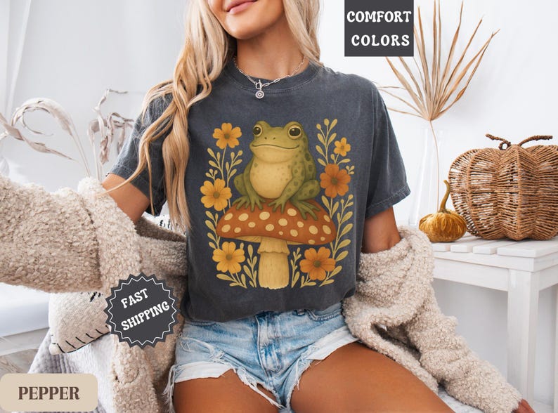 May include: A dark grey t-shirt featuring a graphic of a green frog sitting on a mushroom with yellow flowers. The text "COMFORT COLORS" is in the top right corner of the image.
