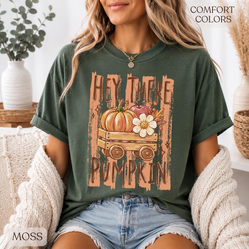 Op de afbeelding: Een donkergroen T-shirt met korte mouwen en een afbeelding van een houten wagen met een pompoen, een appel en een witte bloem. De tekst "HEY THERE PUMPKIN" staat in bruine letters boven en onder de illustratie van de wagen.