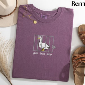 May include: Berry-colored t-shirt with a whimsical embroidered design. The design features a white goose inside a cage with the text "got too silly". A gold necklace and a beige hat are also visible.