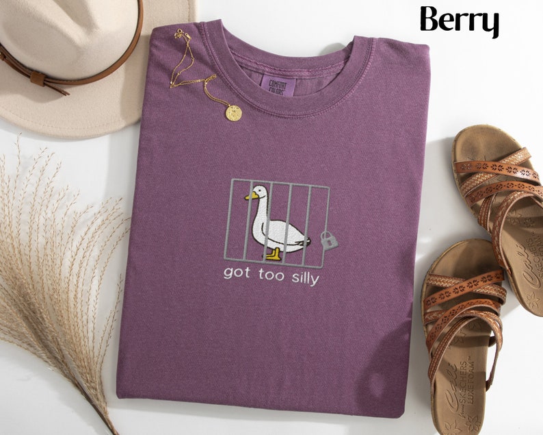 May include: Berry-colored t-shirt with a whimsical embroidered design. The design features a white goose inside a cage with the text "got too silly". A gold necklace and a beige hat are also visible.