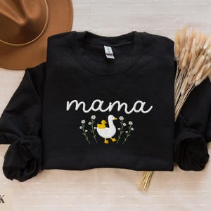 May include: Black sweatshirt with the text "mama" embroidered in white, featuring a white duck with a yellow duckling on its back, surrounded by white flowers.