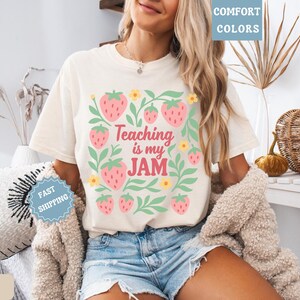 May include: Ivory-colored t-shirt with a strawberry design and the text "Teaching is my JAM." The design includes pink strawberries, green leaves, and yellow flowers. The shirt is a comfort colors style.