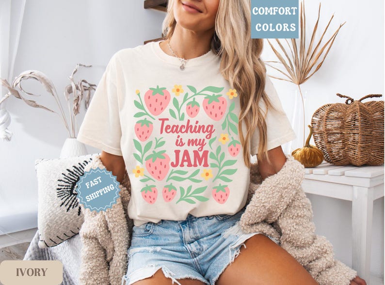 May include: Ivory-colored t-shirt with a strawberry design and the text "Teaching is my JAM." The design includes pink strawberries, green leaves, and yellow flowers. The shirt is a comfort colors style.