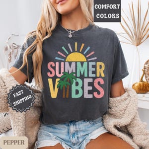 K&ouml;nnte beinhalten: Dunkelgraues T-Shirt mit dem Schriftzug "SUMMER VIBES" in bunten Buchstaben, einer gelben Sonne und Palmen. Das Shirt hat einen Vintage-Look und ist Teil einer Sommerkollektion.