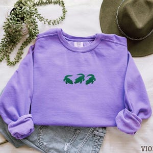 May include: A violet-colored sweatshirt with a crew neck and long sleeves. The front of the shirt features three green alligator designs. The word "VIOLET" is printed in the bottom right corner of the image. The sweatshirt is folded on top of a pair of jeans.