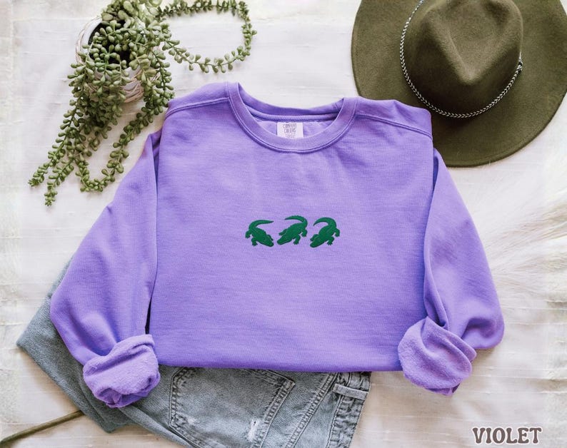 May include: A violet-colored sweatshirt with a crew neck and long sleeves. The front of the shirt features three green alligator designs. The word "VIOLET" is printed in the bottom right corner of the image. The sweatshirt is folded on top of a pair of jeans.