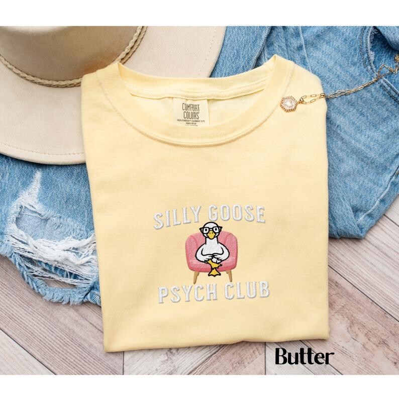 May include: A yellow t-shirt featuring the text "Silly Goose Psych Club" and an embroidered image of a goose wearing glasses and sitting in a pink armchair.