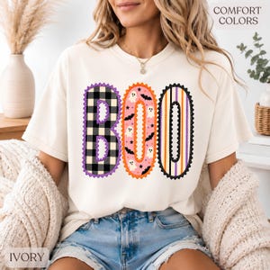 May include: An ivory t-shirt featuring the text BOO in large letters. The B is black and white buffalo plaid, the O is pink with white ghosts and black bats, and the second O has vertical orange, purple, and white stripes. The text COMFORT COLORS is visible.
