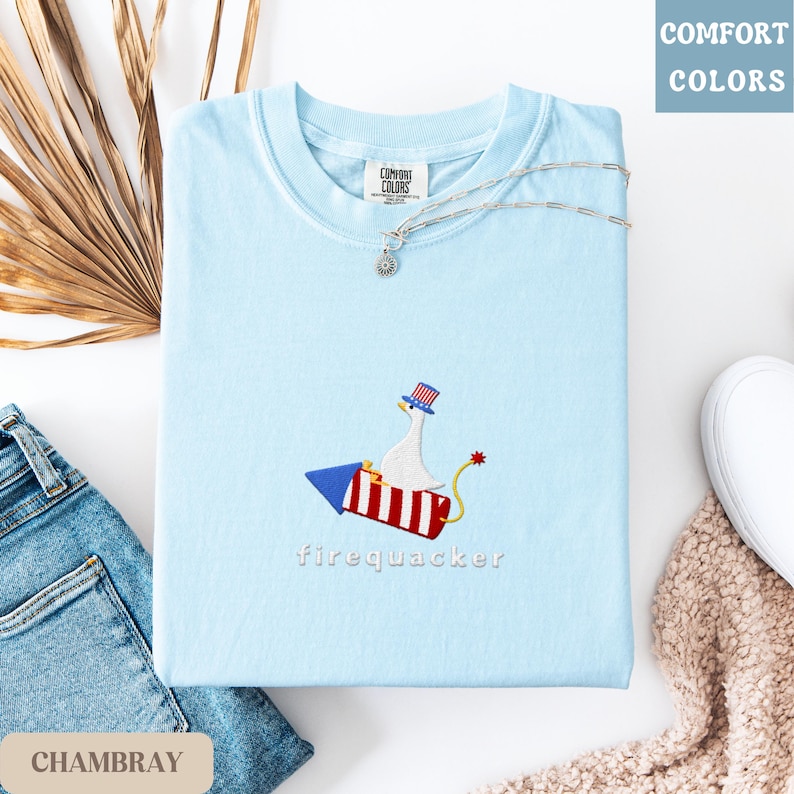K&ouml;nnte beinhalten: Hellblaues T-Shirt mit einem gestickten Design einer Gans auf einer Rakete, darunter das Wort "firequacker". Das Shirt hat einen Rundhalsausschnitt und kurze &Auml;rmel. Eine silberne Halskette ist &uuml;ber dem Shirt drapiert. Die Worte "Comfort Colors" stehen oben rechts.