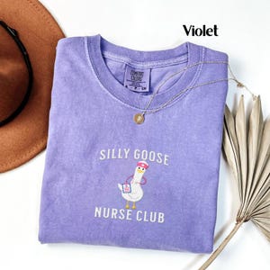 Puede incluir: Una camiseta violeta Comfort Colors con el texto "SILLY GOOSE NURSE CLUB" y una ilustraci&oacute;n de un ganso con gorro de enfermera y bloc de notas. Un collar dorado con un colgante est&aacute; sobre la camiseta.