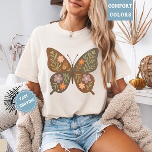 Peut inclure: T-shirt de couleur ivoire avec un motif de papillon marron orn&eacute; de motifs floraux et de feuilles verts, violets et oranges. Le t-shirt porte les mentions "Comfort Colors" et "Fast Shipping".