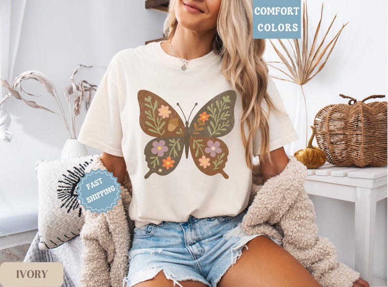 Peut inclure: T-shirt de couleur ivoire avec un motif de papillon marron orn&eacute; de motifs floraux et de feuilles verts, violets et oranges. Le t-shirt porte les mentions "Comfort Colors" et "Fast Shipping".