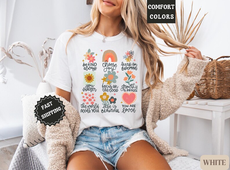 Pu&ograve; includere: Maglietta bianca con un design floreale colorato e affermazioni positive, tra cui "sii sempre gentile", "scegli la gioia", "continua a fiorire", "tu conti", "concentrati sul bene", "la crescita &egrave; un processo", "il mondo ha bisogno di te", "la vita &egrave; bella" e "sei amato".