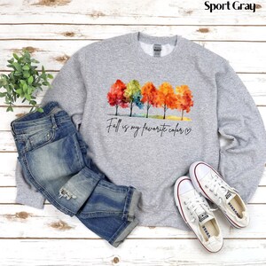 May include: A gray sweatshirt with a watercolor design of colorful fall trees and the text "Fall is my favorite color."  The sweatshirt is paired with blue jeans and white sneakers.