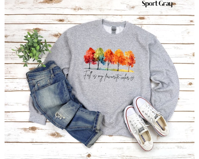 May include: A gray sweatshirt with a watercolor design of colorful fall trees and the text "Fall is my favorite color."  The sweatshirt is paired with blue jeans and white sneakers.