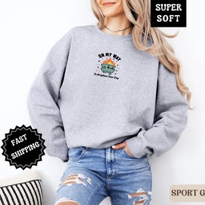 Pu&ograve; includere: Felpa grigio sport con una grafica di un bidone della spazzatura in fiamme, con la scritta "ON MY WAY To Brighten Your Day". La felpa &egrave; realizzata in un materiale morbido.