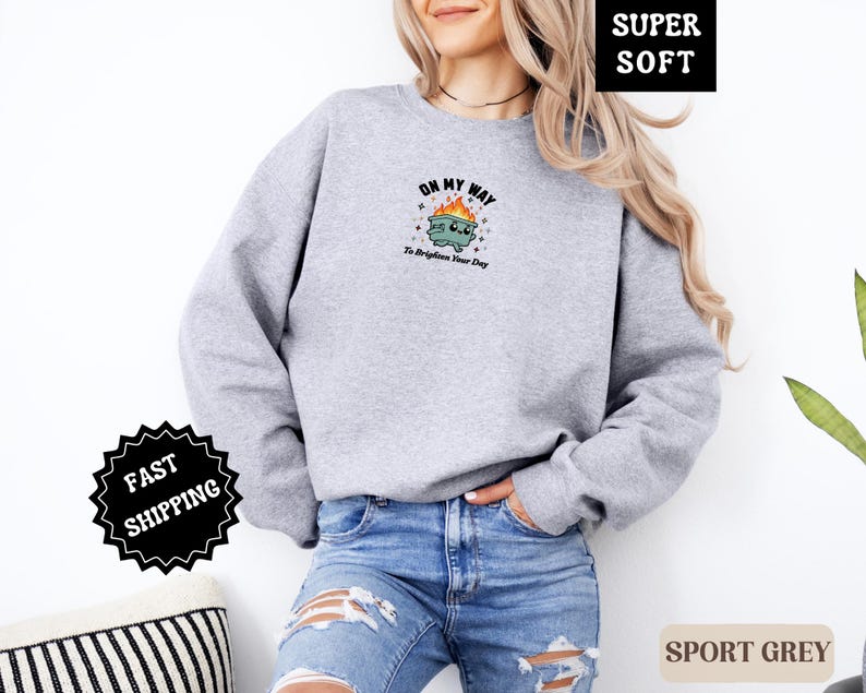 Pu&ograve; includere: Felpa grigio sport con una grafica di un bidone della spazzatura in fiamme, con la scritta "ON MY WAY To Brighten Your Day". La felpa &egrave; realizzata in un materiale morbido.