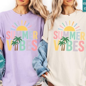 K&ouml;nnte beinhalten: Zwei T-Shirts, eines in Orchidee und eines in Elfenbein, mit dem Text "SUMMER VIBES" in bunten Buchstaben, einer Sonne und Palmen. Die Shirts haben eine lockere Passform, perfekt f&uuml;r den Sommer.