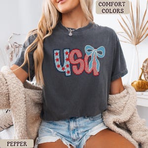 Op de afbeelding: Donkergrijs t-shirt met het woord "USA" in rood, wit en blauw, met een strik. Het shirt heeft een vintage uitstraling. De tekst "COMFORT COLORS" staat rechtsboven en "PEPPER" linksonder.