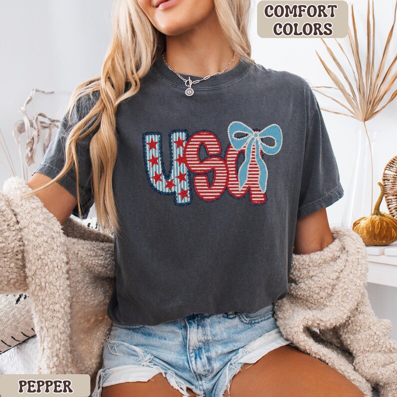 Op de afbeelding: Donkergrijs t-shirt met het woord "USA" in rood, wit en blauw, met een strik. Het shirt heeft een vintage uitstraling. De tekst "COMFORT COLORS" staat rechtsboven en "PEPPER" linksonder.