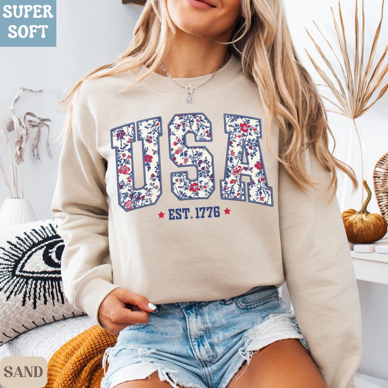 Peut inclure: Sweat-shirt couleur sable avec le mot "USA" en motif floral, encadr&eacute; de bleu marine. Le texte "EST. 1776" est en dessous avec des &eacute;toiles rouges. Le sweat-shirt a des manches longues et un col rond.