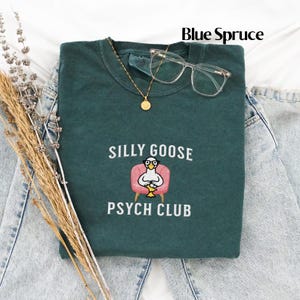 May include: A dark green t-shirt featuring the text "Silly Goose Psych Club" and an embroidered image of a goose wearing glasses and sitting in a pink armchair.