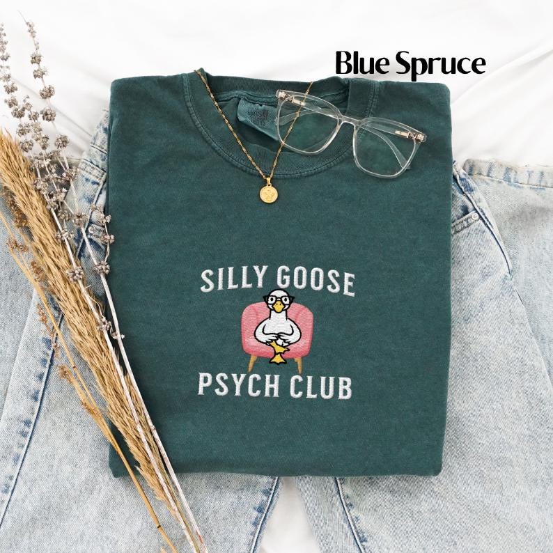 May include: A dark green t-shirt featuring the text "Silly Goose Psych Club" and an embroidered image of a goose wearing glasses and sitting in a pink armchair.