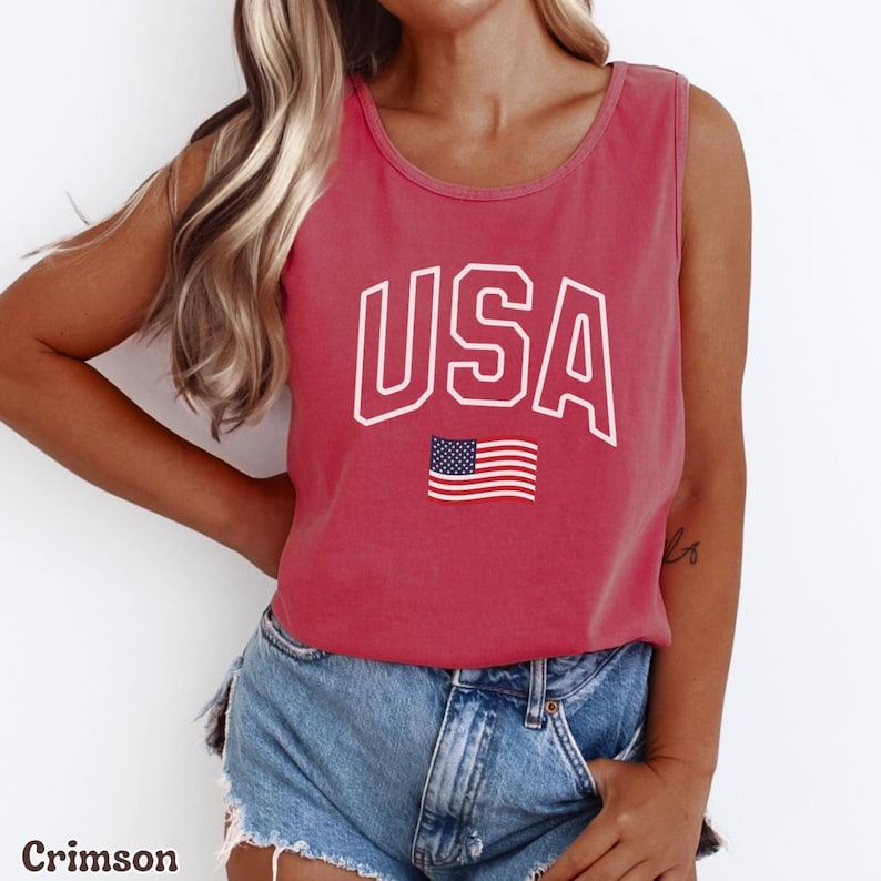 Peut inclure: D&eacute;bardeur cramoisi avec le mot "USA" en lettres blanches et un petit drapeau am&eacute;ricain. Le d&eacute;bardeur est sans manches et a une encolure arrondie. Le mod&egrave;le porte un short en jean.
