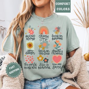 Pu&ograve; includere: Una maglietta blu chiaro con una stampa grafica colorata che presenta fiori, cuori e messaggi ispiratori. Il testo include "be kind always", "choose joy", "keep on blooming", "you matter", "focus on the good", "growth is a process", "the world needs you", "life is beautiful" e "you are loved".