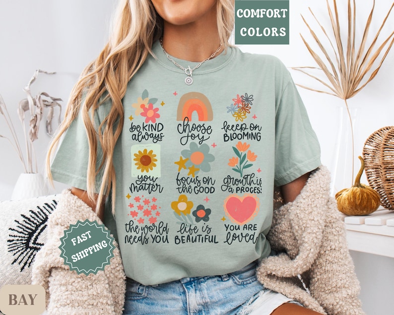 Pu&ograve; includere: Una maglietta blu chiaro con una stampa grafica colorata che presenta fiori, cuori e messaggi ispiratori. Il testo include "be kind always", "choose joy", "keep on blooming", "you matter", "focus on the good", "growth is a process", "the world needs you", "life is beautiful" e "you are loved".