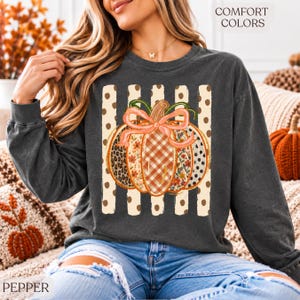 May include: A charcoal grey long-sleeve sweatshirt featuring a graphic of a pumpkin with a pink bow. The pumpkin has sections with leopard print, plaid, and floral patterns. Behind the pumpkin are vertical beige stripes with brown polka dots. The text COMFORT COLORS is visible in the top right corner.