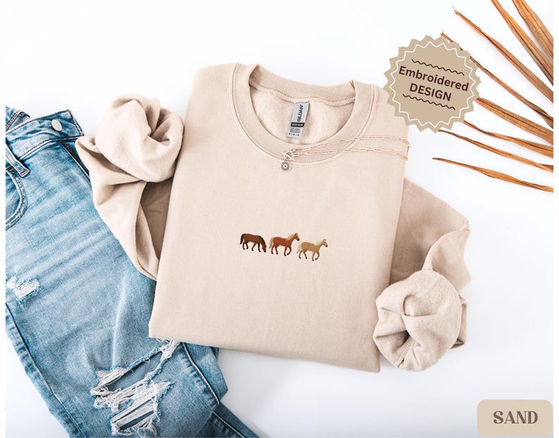 K&ouml;nnte beinhalten: Beigefarbenes Sweatshirt mit einer Stickerei von drei Pferden in Braun- und Beiget&ouml;nen. Das Sweatshirt liegt neben einer zerrissenen blauen Jeans. Der Text "Embroidered DESIGN" befindet sich auf einem dekorativen Etikett.