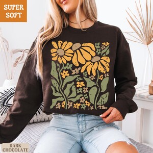 May include: Dark chocolate-colored sweatshirt with a floral design. The graphic features large yellow flowers, green leaves, and smaller flowers. The words "SUPER SOFT" and "DARK CHOCOLATE" are printed on the image.