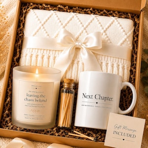 May include: A gift box containing a white knit blanket tied with a satin ribbon, a frosted glass candle labeled 'leaving the chaos behind', a small glass vial of matches, a white mug with 'Next Chapter.' printed on it, and a gift message card.