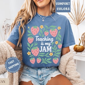 May include: Blue jean short-sleeved t-shirt with a strawberry and floral design. The shirt features the text "Teaching is my JAM" in a playful font. The design includes pink strawberries and green leaves.