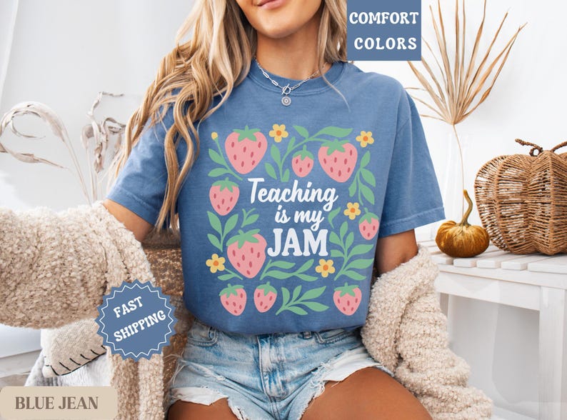 May include: Blue jean short-sleeved t-shirt with a strawberry and floral design. The shirt features the text "Teaching is my JAM" in a playful font. The design includes pink strawberries and green leaves.