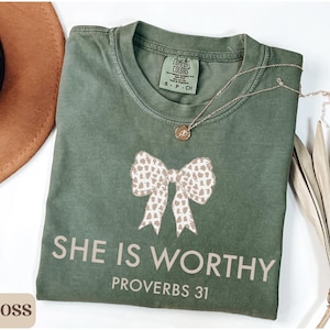 May include: A moss green t-shirt with a bow graphic and the text "SHE IS WORTHY PROVERBS 31". A gold necklace with a pendant is on the shirt. A brown hat is in the background.