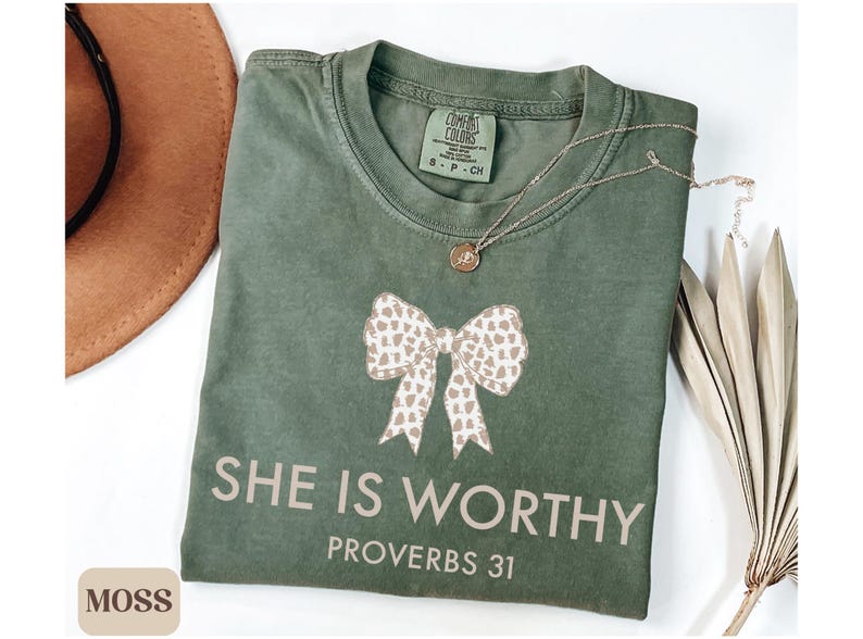 May include: A moss green t-shirt with a bow graphic and the text "SHE IS WORTHY PROVERBS 31". A gold necklace with a pendant is on the shirt. A brown hat is in the background.