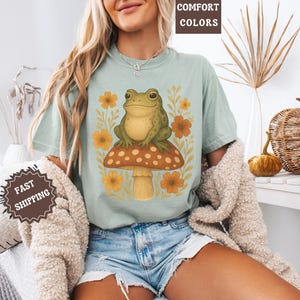 May include: A light blue t-shirt featuring a graphic of a green frog sitting on a mushroom with yellow and orange flowers. The text "COMFORT COLORS" is visible in the top right corner of the image.