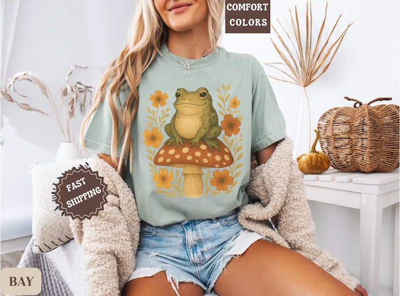 May include: A light blue t-shirt featuring a graphic of a green frog sitting on a mushroom with yellow and orange flowers. The text "COMFORT COLORS" is visible in the top right corner of the image.