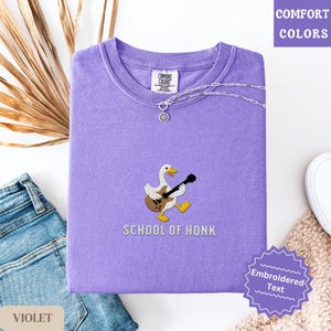 May include: Violet Comfort Colors sweatshirt with a crew neck. The shirt features an embroidered design of a goose playing a guitar, with the text "SCHOOL OF HONK" below. A silver necklace with a pendant is draped on the shirt.