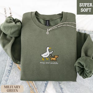 May include: Military green sweatshirt with the text "wag and waddle" and a graphic of a duck walking a dog. The sweatshirt is labeled "Super Soft" and is displayed on a light blue denim surface.