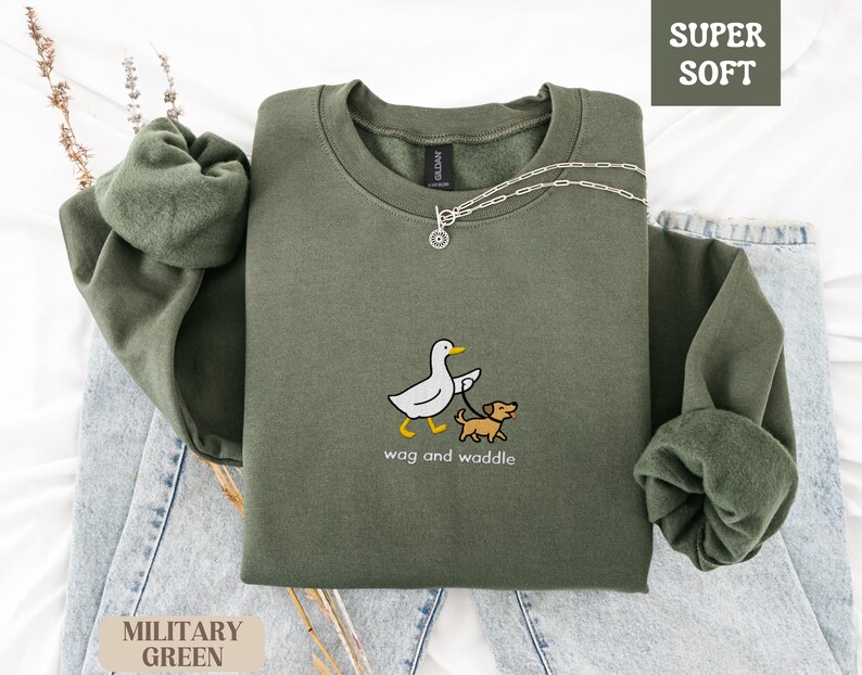 May include: Military green sweatshirt with the text "wag and waddle" and a graphic of a duck walking a dog. The sweatshirt is labeled "Super Soft" and is displayed on a light blue denim surface.