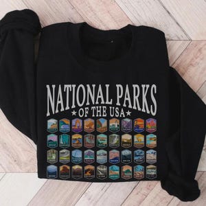 May include: Black t-shirt with the text "National Parks of the USA" in white lettering. The shirt features a grid of colorful national park logos.