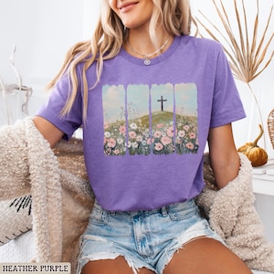 May include: Heather purple t-shirt featuring a graphic of a cross on a hill with flowers. The design is framed by vertical rectangular shapes. The shirt is short-sleeved and made of a soft material. The text "HEATHER PURPLE" is at the bottom of the image.