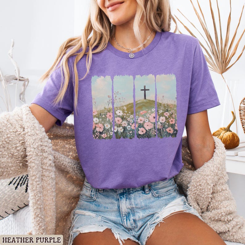 May include: Heather purple t-shirt featuring a graphic of a cross on a hill with flowers. The design is framed by vertical rectangular shapes. The shirt is short-sleeved and made of a soft material. The text "HEATHER PURPLE" is at the bottom of the image.