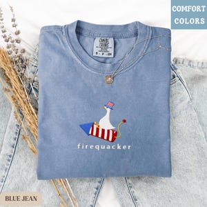 K&ouml;nnte beinhalten: Ein blaues Comfort Colors T-Shirt mit einem gestickten Design. Das Design zeigt eine wei&szlig;e Gans mit patriotischem Hut, die auf einer rot-wei&szlig;-blauen Rakete reitet, mit dem Wort "firequacker" darunter. Eine silberne Halskette liegt auf dem Shirt.