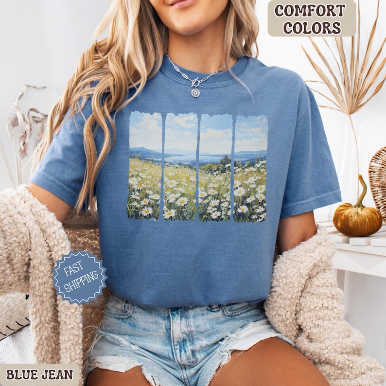 May include: Blue jean colored t-shirt with a landscape design. The design features a field of white daisies, a blue sky, and a body of water. The words "Comfort Colors" are printed on the top right.