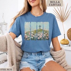 May include: Steel blue t-shirt with a graphic of a cross on a hill surrounded by flowers. The text "BELLA CANVAS" is in the upper right corner. The shirt is a short-sleeved crew neck.