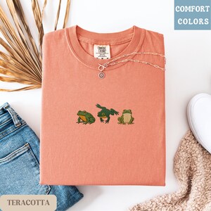 May include: Terracotta-colored t-shirt with three embroidered frog designs. A silver necklace with a sun pendant rests on the shirt. The shirt is folded with a pair of jeans and a white shoe in the background. The text "COMFORT COLORS" is in the upper right corner.